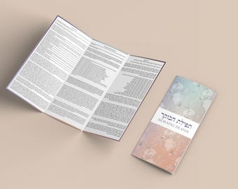 Könnte beinhalten: Eine Broschüre mit dem Text "Morning Prayer" auf Englisch und Hebräisch. Die Broschüre ist geöffnet und zeigt textgefüllte Seiten. Daneben liegt eine geschlossene Broschüre mit einem floralen Design in Blau-, Pfirsich- und Weißtönen.