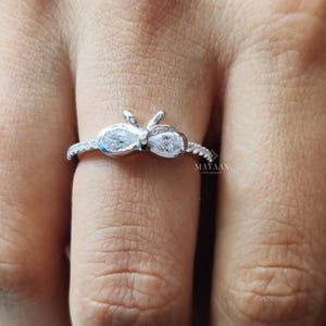 以下が含まれることがあります： 蝶のデザインのシルバーリング。中央にペアシェイプの石が2つ、バンドに沿って小さな石が並んでいます。指にはめられており、繊細なディテールと輝きが際立っています。ブランドはMAYAANです。