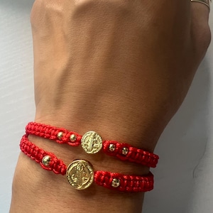 Könnte beinhalten: Zwei rote geflochtene Armbänder mit goldfarbenen Akzenten und runden Medaillons. Die Medaillons weisen ein detailliertes Design auf. Die Armbänder werden an einem Handgelenk getragen.