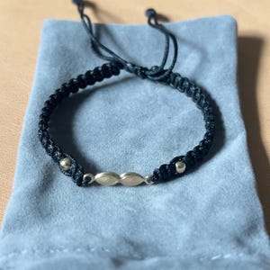 Peut inclure: Un bracelet en cordon noir avec un design en macramé tressé, deux petites perles métalliques et une breloque centrale horizontale dorée en forme de deux maillons ovales connectés. Le bracelet possède des cordons ajustables.