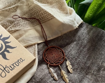 May include: A brown dreamcatcher with three gold-colored feathers hangs against a neutral-toned fabric. A small cardboard box with the word "Aurora" is visible. The dreamcatcher is a circular woven design.