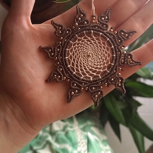 Könnte beinhalten: Eine handgefertigte Makramee-Anhänger-Halskette mit einem Sonnenstrahlen-Design. Der Anhänger ist braun mit einem cremefarbenen Zentrum und goldfarbenen Perlen. Die Halskette wird in einer Hand gehalten.