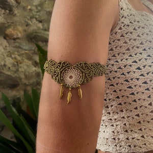 Pode incluir: Uma pulseira de macramé verde com um amuleto de apanhador de sonhos branco e detalhes de penas douradas.