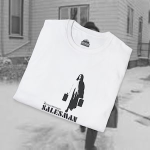 Puede incluir: Camiseta blanca con un gráfico en blanco y negro de una persona con maletines y un halo. El texto "THE MAYSLES BROTHERS SALESMAN" está impreso debajo. La camiseta tiene cuello redondo y una etiqueta en el cuello. Diseño monocromático.