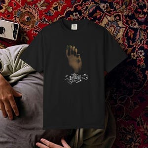 May include: Black t-shirt with a graphic of a hand with green nail polish. The graphic is above the text "PIO LESINSKI" in a stylized font. The shirt has a crew neck and is likely made of cotton.