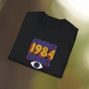 May include: Black t-shirt featuring the text "GEORGE ORWELL 1984" in a retro, layered design with red, yellow, and blue colors. Below the text is a white eye with a blue iris. The shirt is folded.