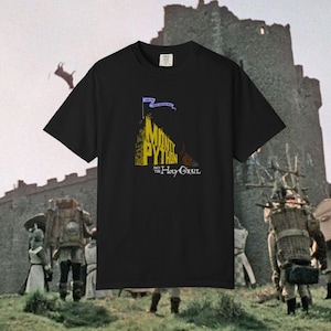 May include: Black t-shirt with the words "Monty Python and the Holy Grail" in yellow and blue, with a castle illustration and banner. The shirt is set against a backdrop of a medieval castle and figures, evoking a historical theme.