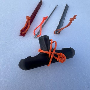 May include: Four tent pegs with orange ropes. Three pegs are metal and one is black plastic. The pegs are on a white snowy surface.