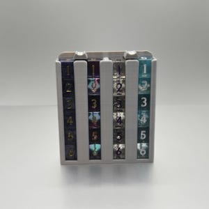 May include: A white plastic dice tower with four columns. Each column holds six dice with different colors and numbers. The dice are numbered 1 through 6.