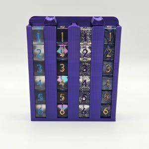 May include: A purple plastic storage rack holding four vertical columns of six-sided dice. Each column contains six dice numbered one through six. The dice feature various finishes including translucent blue, iridescent, silver glitter, and marbled grey.