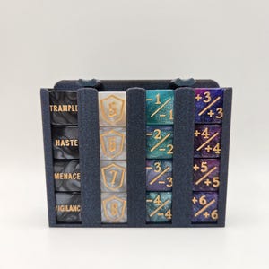 May include: A dark blue dice holder with multiple dice. The dice are in various colors, including black, white, teal, and purple. The dice have gold numbers and symbols. The holder has text that reads "TRAMPLE", "HASTE", "MENACE", and "VIGILANCE".