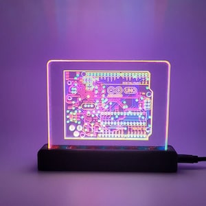 May include: A clear acrylic lamp base with a black base and a circuit board design inside. The circuit board is lit up with rainbow colors and has the text "Arduino UNO" printed on it.