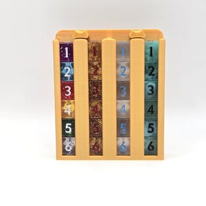 May include: A wooden dice tower with three columns of dice. Each column has six dice with numbers 1-6. The dice are various colors, including blue, green, and gold. The tower is a light brown color.