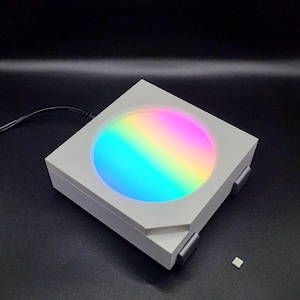 May include: A white square box with a round, colorful light inside. The light is a gradient of colors, starting with blue and transitioning to pink. The box has a small, white square on the side.