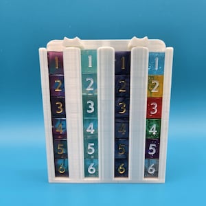 May include: A white 3D-printed dice holder with six columns. Each column holds six multi-colored dice, numbered 1-6 in gold. The dice colors include purple, teal, blue, and red. The holder has a popcorn box design.