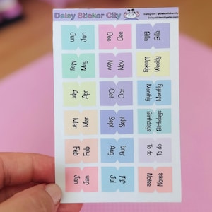 May include: A sheet of colorful planner stickers with the text "Delay Sticker City" at the top. The stickers are rectangular with rounded corners and feature labels for months, weekly, monthly, and to-do lists. The background is a soft pink.