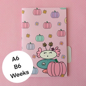 May include: A pink notebook with a cute cartoon character holding a pumpkin. The notebook has a repeating pattern of pumpkins and flowers. The text "A6 B6 Weeks" is printed on the front of the notebook.
