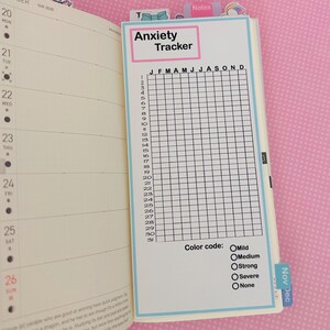 May include: A planner insert with the text "Anxiety Tracker" in a pink and blue frame. The insert features a grid for tracking anxiety levels throughout the year, with a color code for different intensities. The planner is open to the October 2025 page.