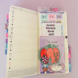 May include: Open planner with August calendar, stickers, and a pocket featuring a pumpkin illustration with flowers and the text "Be around re". Includes stickers from "Delay Sticker City" and month labels.