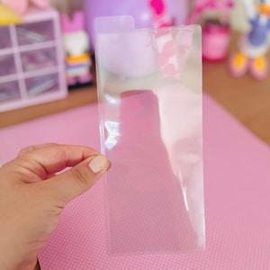 May include: A clear, rectangular plastic sleeve with a small tab at the top is held in a hand. The sleeve is transparent, allowing a pink surface with white polka dots to be seen through it. The background includes pink and purple decorative items.