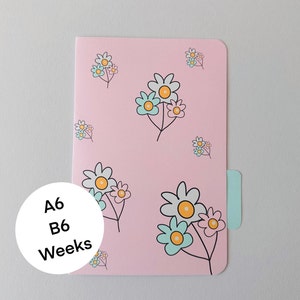 May include: A pink weekly planner with a floral pattern. The planner has a tab on the side and the text "A6 B6 Weeks" on the front.