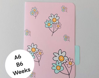 May include: A pink weekly planner with a floral pattern. The planner has a tab on the side and the text "A6 B6 Weeks" on the front.