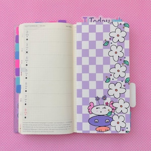 May include: A purple and white checkered planner with a floral border and a cartoon character on the right page. The planner has the word "SEPTEMBER" and the numbers "2" through "8" on the left page.