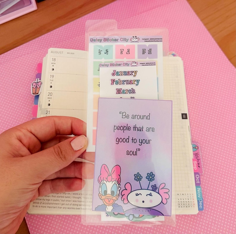 May include: A planner with a clear pocket holding stickers and a card with the text "Be around people that are good to your soul." The planner has monthly tabs and a calendar.