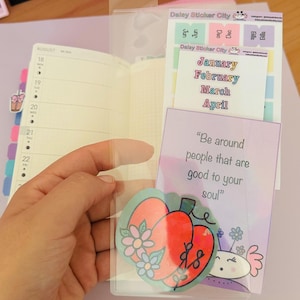 May include: A clear plastic planner pocket holding stickers and a card. The stickers are labeled "Jan, Feb, Mar, April" and "Bills". The card reads "Be around people that are good to your soul". A planner is visible.