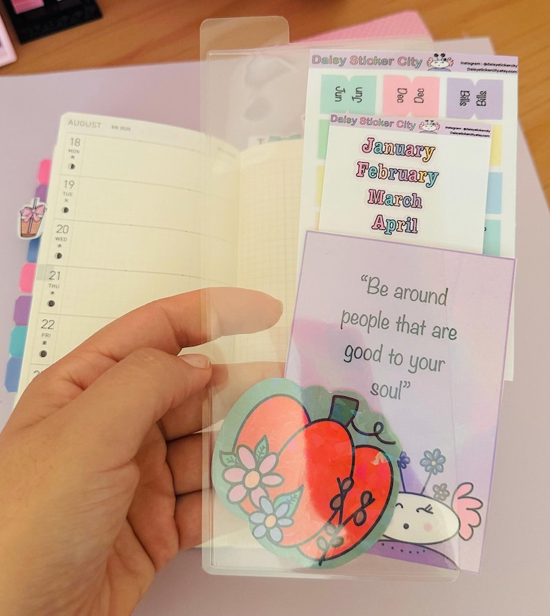 May include: A clear plastic planner pocket holding stickers and a card. The stickers are labeled "Jan, Feb, Mar, April" and "Bills". The card reads "Be around people that are good to your soul". A planner is visible.