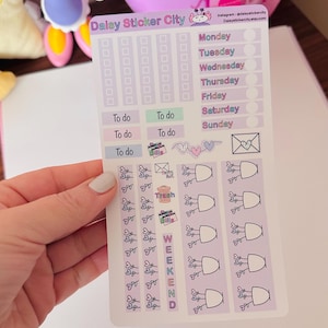 May include: A planner sticker sheet with a light purple background. The sheet includes boxes for each day of the week, "To do" labels, and various small illustrations of hearts, envelopes, and potted plants. The text "Daisy Sticker City" is visible.