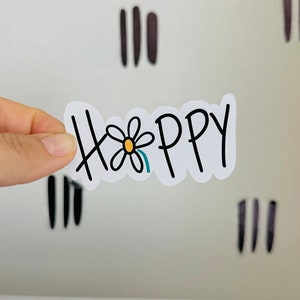 May include: A white sticker with the word "Happy" in black handwritten font, with a daisy replacing the "o". The sticker is held against a white background with vertical black and brown lines.