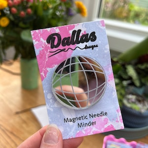 Op de afbeelding: Een witte kaart met roze en blauwe marmering. De kaart heeft de tekst "Dallas Designs" in zwarte cursieve letters. Een zilveren, ronde, magnetische naaldhouder met een rasterpatroon bevindt zich op de kaart. De tekst "Magnetic Needle Minder" is onder de houder gedrukt.