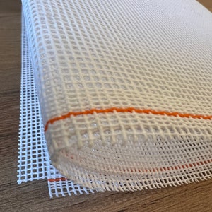 Op de afbeelding: Close-up van een wit tapijtcanvas met een rasterpatroon. De rand van het canvas is zichtbaar, met een oranje draaddetail. Het canvas is opgerold, waardoor de textuur en constructie van het materiaal zichtbaar worden.