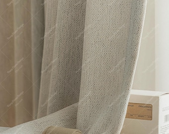 May include: A light beige curtain with a textured, woven fabric finish hangs in soft vertical folds. A small, cylindrical, tan-colored spool rests on the fabric. A white cardboard box with the text "sunset FRAGRANCE" and "AOOYVUE" is visible.