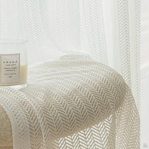 Pode incluir: Uma vela acesa em um frasco de vidro transparente com o texto "ARAMA" está sobre uma manta bege texturizada. A manta tem um padrão de espinha de peixe. Cortinas brancas com um padrão semelhante estão ao fundo.