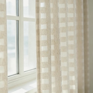 Puede incluir: Cortinas beige y blancas estampadas colgadas frente a una ventana. Las cortinas tienen un aspecto texturizado con un patrón de cuadrícula de cuadrados blancos sobre un fondo beige. El marco de la ventana es blanco.