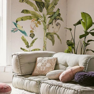 May include: A light green modular sofa with multiple cushions and pillows. The sofa is decorated with a tropical theme featuring a banana tree, a sloth, a toucan, and a parrot. The sofa is in a room with a white wall and a white rug.