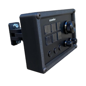 Op de afbeelding: Zwart bedieningspaneel met knoppen, knoppen en de tekst "soomfon". Het paneel heeft een gestructureerd oppervlak en is gemonteerd op een zwarte beugel. Het heeft heldere, rechthoekige knoppen en draaiknoppen.