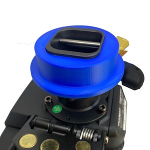 Può includere: Una base per volante da corsa nera con un prominente adattatore circolare blu montato sulla parte superiore. L'unità include un meccanismo di sgancio rapido in metallo nero con tre punti di contatto circolari dorati e una cerniera a molla.