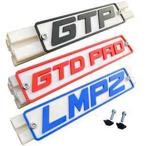 Op de afbeelding: Drie rechthoekige platen met de tekst GTP, GTD PRO en LMP2 in verhoogde zwarte, rode en blauwe letters op een witte achtergrond. Elke plaat is gemonteerd op een witte gegroefde basis met zilverkleurige metalen bouten in de hoeken.