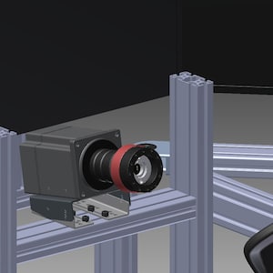 May include: A gray industrial camera with a black lens and a red ring mount, secured to a silver aluminum extrusion frame. The camera is positioned horizontally, featuring a metallic mounting bracket attached to the structural metal beams.