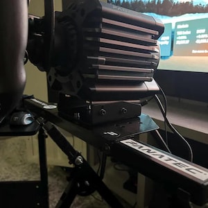 May include: A black Fanatec direct drive racing wheel base mounted on a metal frame. The unit features a ribbed cooling housing and is attached to a black support structure with the Fanatec logo clearly visible on the frame arm and mounting plate.