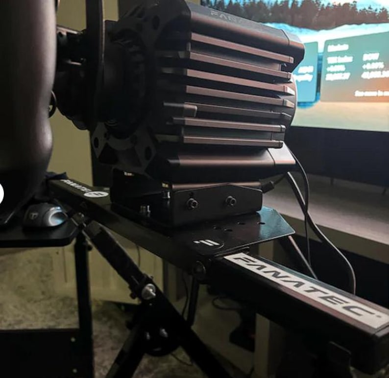 May include: A black Fanatec direct drive racing wheel base mounted on a metal frame. The unit features a ribbed cooling housing and is attached to a black support structure with the Fanatec logo clearly visible on the frame arm and mounting plate.