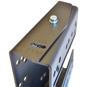 May include: A matte black metal bracket featuring a U-shaped profile with multiple circular mounting holes along the sides. The top surface includes a central silver bolt and a rectangular slot with etched alignment markings for precise positioning.
