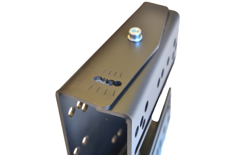 May include: A matte black metal bracket featuring a U-shaped profile with multiple circular mounting holes along the sides. The top surface includes a central silver bolt and a rectangular slot with etched alignment markings for precise positioning.