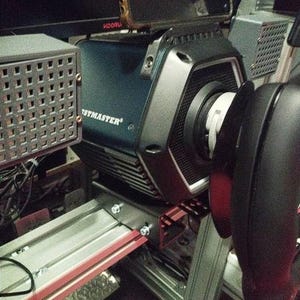 May include: A dark blue Thrustmaster racing wheel base featuring a hexagonal front housing with a black textured finish, mounted on a silver metal frame with visible bolts, positioned next to a gray perforated metal speaker enclosure.