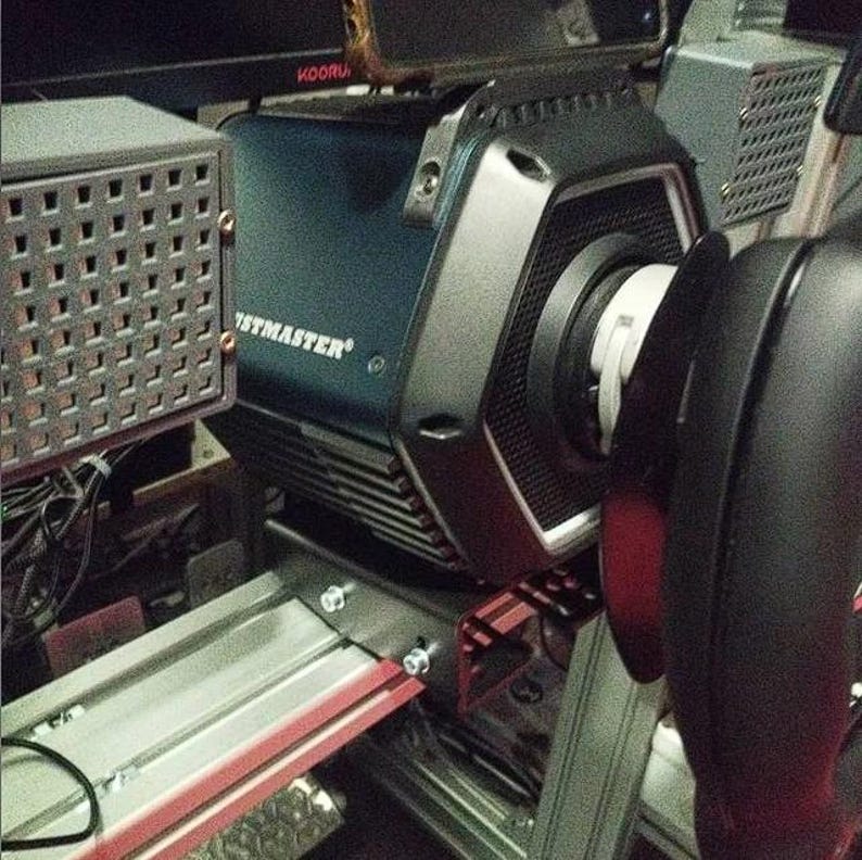 May include: A dark blue Thrustmaster racing wheel base featuring a hexagonal front housing with a black textured finish, mounted on a silver metal frame with visible bolts, positioned next to a gray perforated metal speaker enclosure.