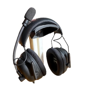 May include: Black over-ear headphones featuring a padded headband, thick fabric ear cushions, and a flexible boom microphone with a foam windscreen. The headset includes thin metal wire supports connecting the ear cups to the adjustable headband.