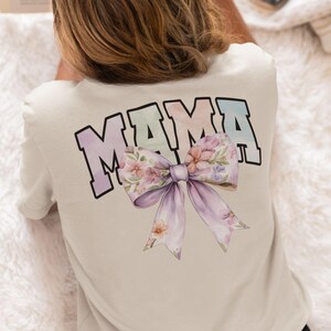Peut inclure: Un t-shirt blanc avec le mot "MAMA" imprim&eacute; en couleurs arc-en-ciel avec un n&oelig;ud floral en dessous.